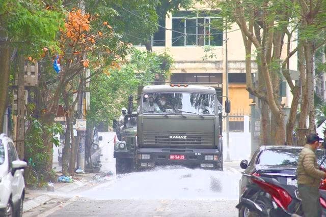 A green Kamaz truck spraying water down a tree-lined street, a parked car and a scooter in the foreground